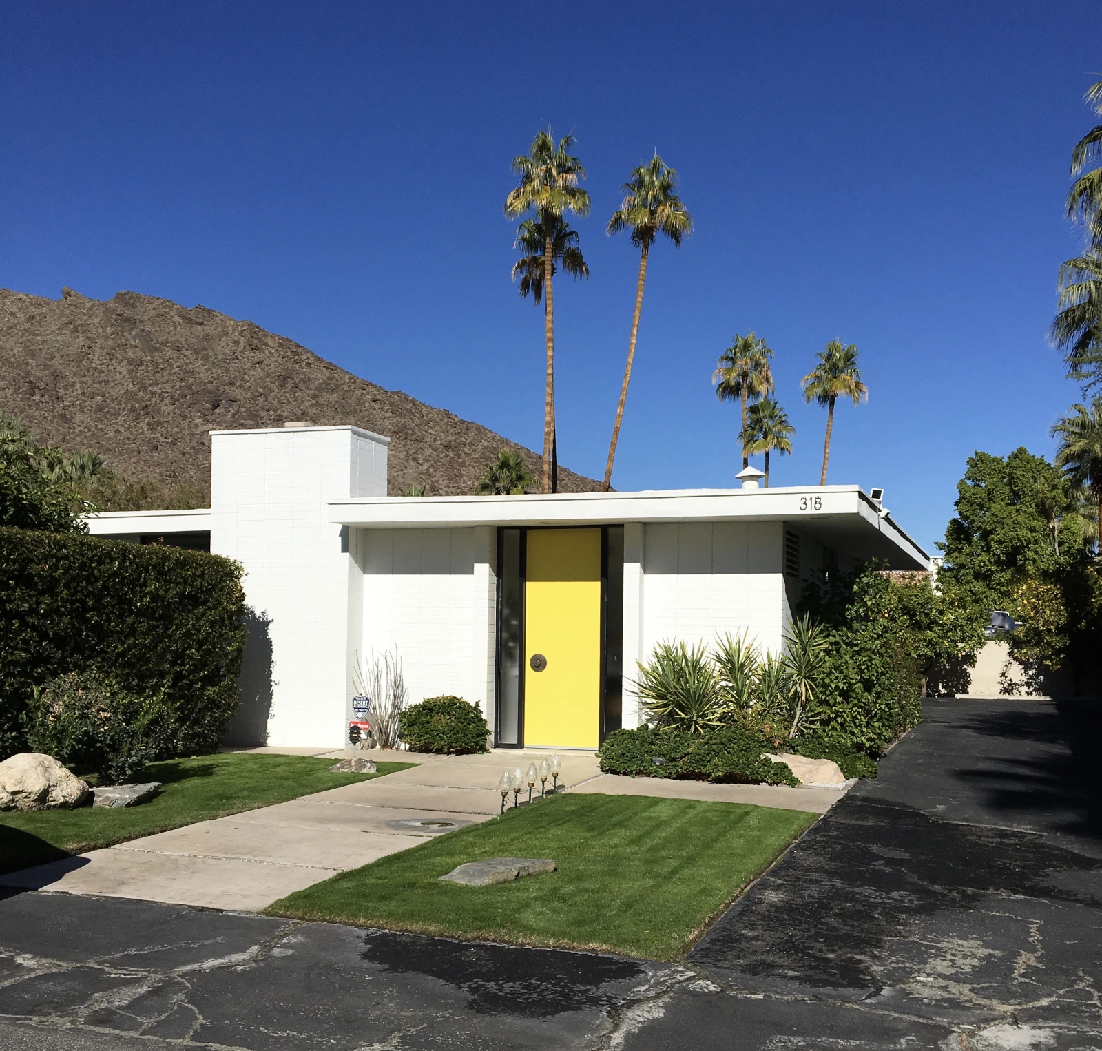 Front view of a small midcentury modern home in Palm Springs with a yellow door, palm trees, and mountains in the distance